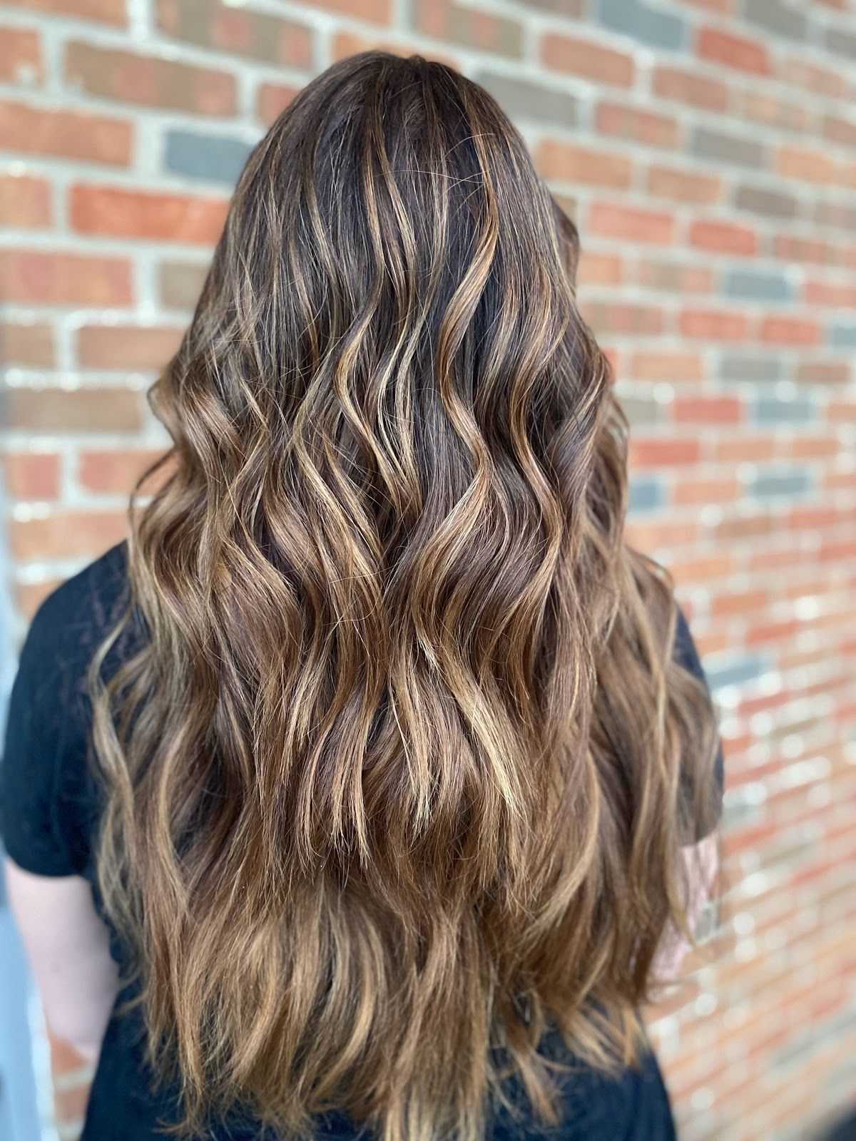 Back view of a person with long, wavy, brown highlighted hair against a brick wall background.